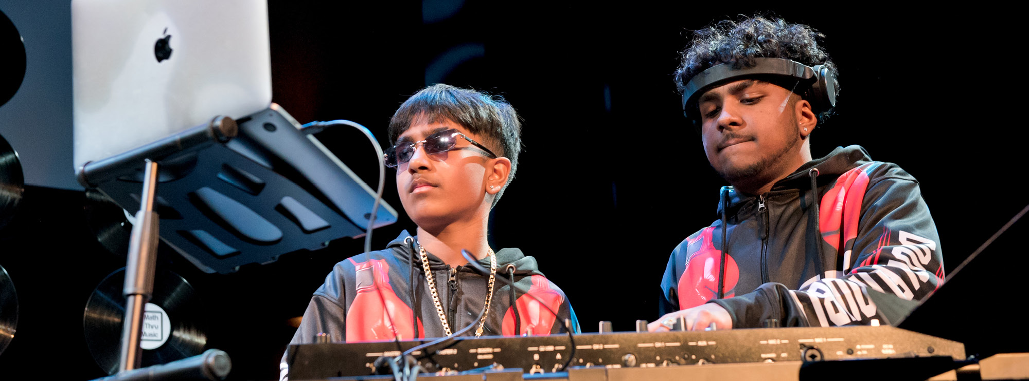 Two young DJs are performing on stage. They are both wearing matching hoodies with bold red and black designs. One DJ is wearing sunglasses and a gold chain, while the other wears headphones. In front of them is a setup with a laptop, mixer, and various controls.