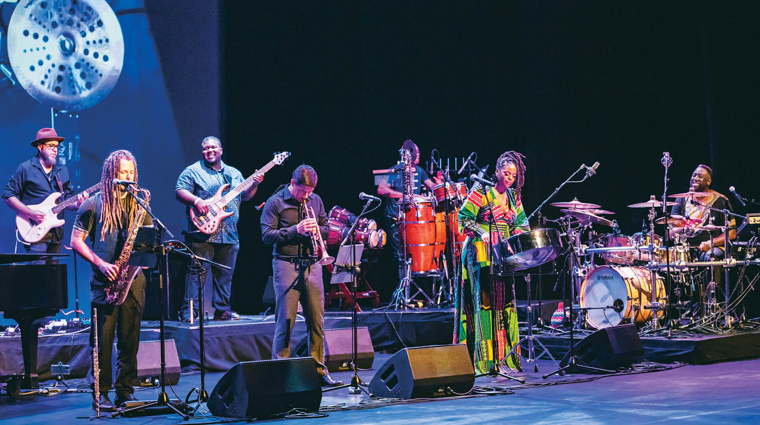 A diverse band performing on stage, featuring musicians playing saxophone, trumpet, electric guitar, bass, steelpan, and drums.