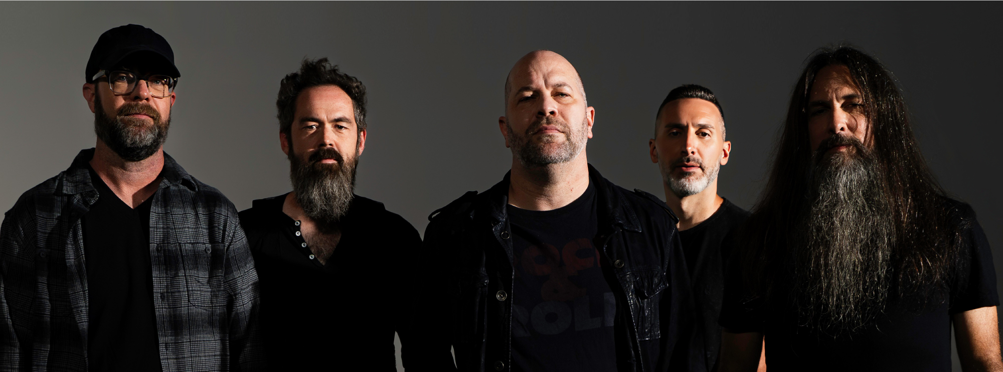 Five men from a rock band standing together against a dark background, wearing black and serious expressions.