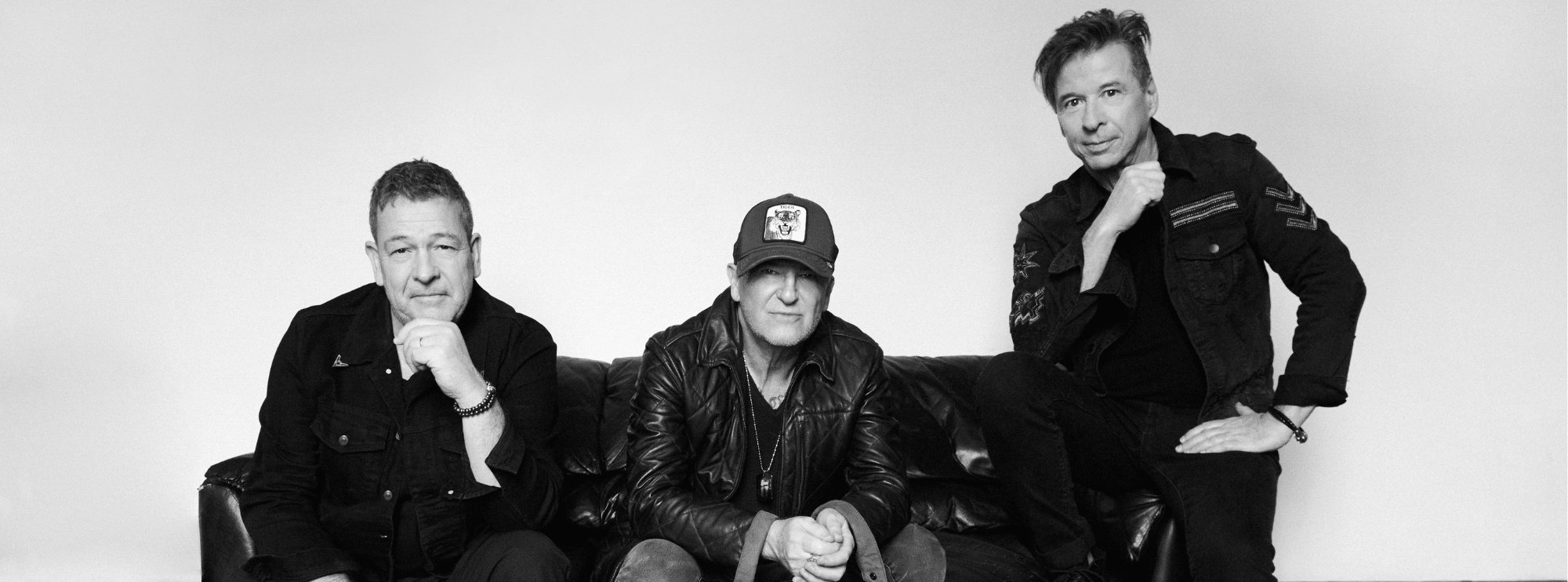 A black and white image of the band Glass Tiger. Three men sit on a black leather couch and look towards the camera. The man most right sits on the couch armchair with his left hand rested on his leg.