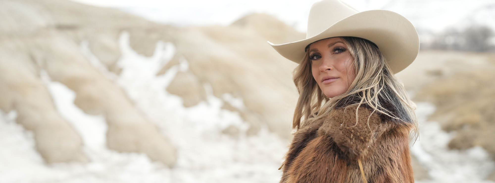 Woman in a cowboy hat and fur coat standing in a snowy, mountainous landscape, looking over her shoulder.