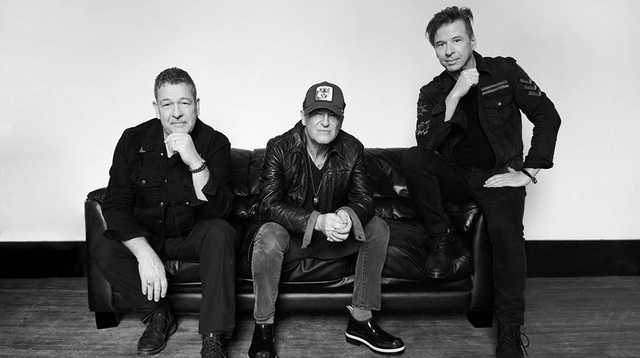 A black and white image of the band Glass Tiger. Three men sit on a black leather couch and look towards the camera. The man most right sits on the couch armchair with his left hand rested on his leg.