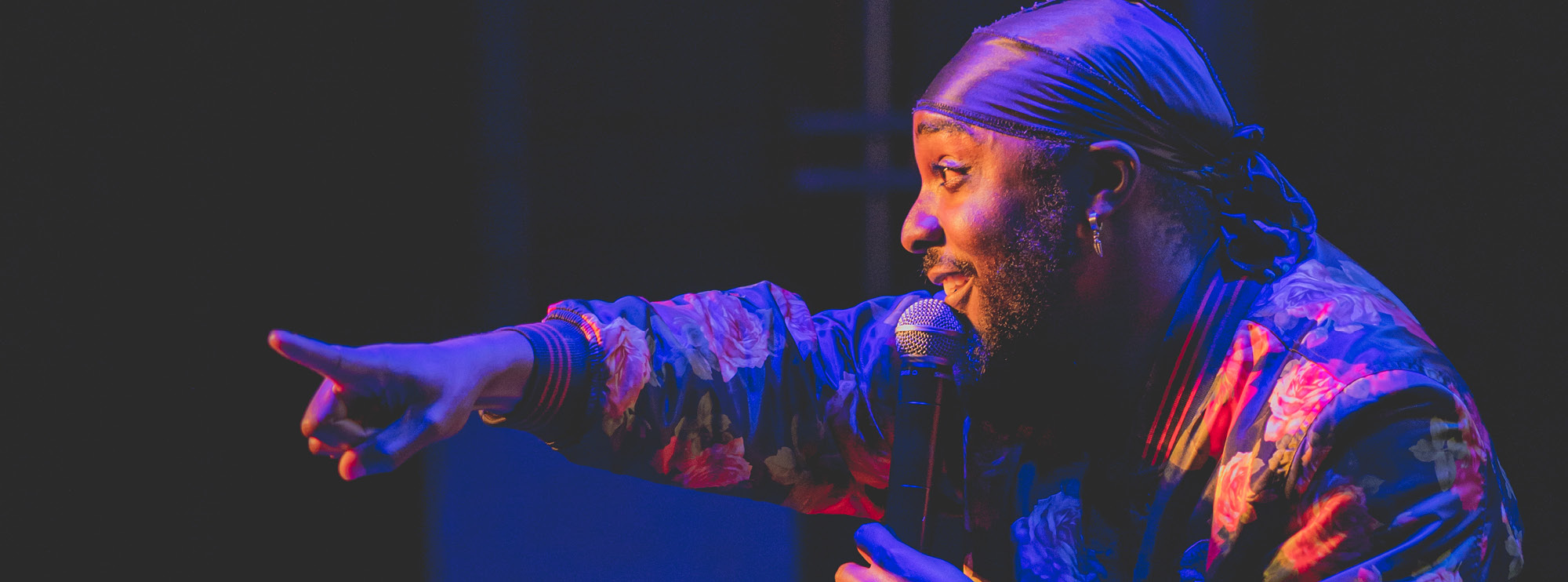 Black male singer with dreadlocks performing against a black backdrop, hand on microphone. 