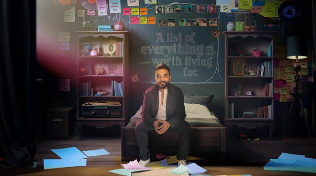 Image of a man seated at the end of a bed with his hands resting in his lap. He has warm-toned skin, a beard, and black hair. Behind him stretches a wall adorned with sticky notes and photographs. In the heart of this collage, text reads: "a list of everything worth living for." Flanking the bed, two tall bookshelves brim with books, framing the scene. Coloured papers scatter the floor.