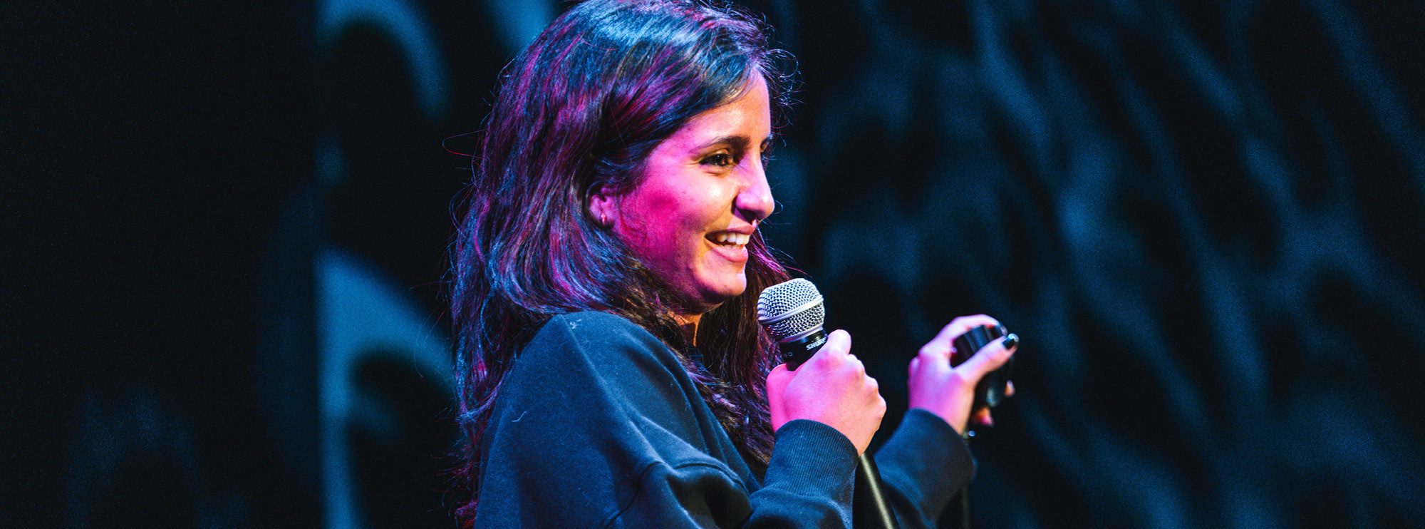 A female comedian is performing on stage, holding a microphone in her right hand and gesturing with her left. Her long dark hair cascades over her shoulders, and she is wearing a black sweater. The stage lighting casts a vibrant mix of pink and blue hues on her face, creating a dynamic and engaging atmosphere. 