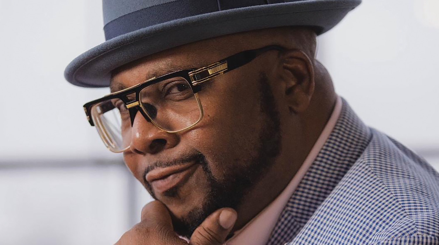 A close-up portrait of a man exudes confidence and style. He is wearing a grey fedora, large glasses with a distinct frame, and a checkered shirt. His expression is composed, with a slight smile and a thoughtful look in his eyes.