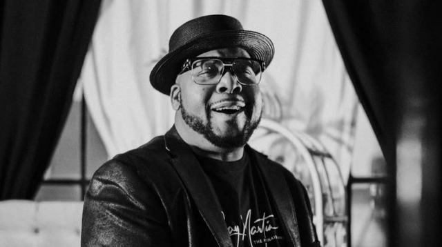 A black and white image of comedian Jay Martin, sporting glasses and a black hat with a flat top and narrow rim. His eyes narrow as a wide smile crosses his face. 