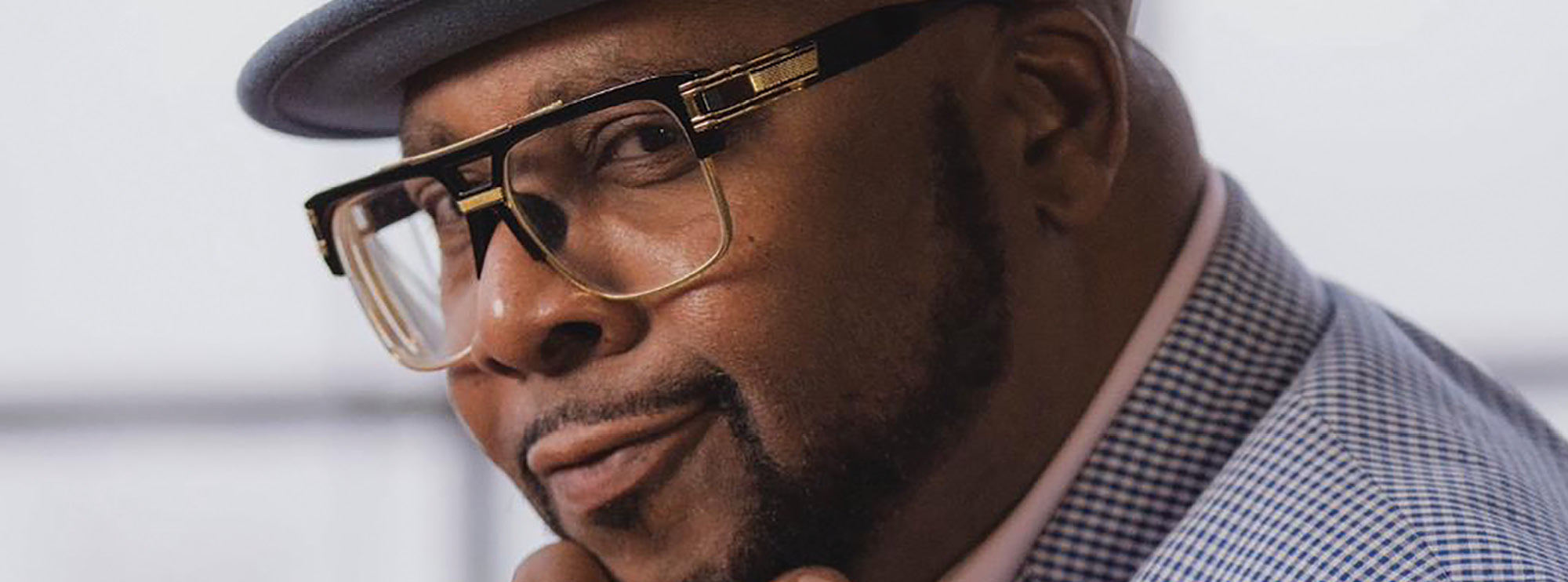 A close-up portrait of a man exudes confidence and style. He is wearing a grey fedora, large glasses with a distinct frame, and a checkered shirt. His expression is composed, with a slight smile and a thoughtful look in his eyes.