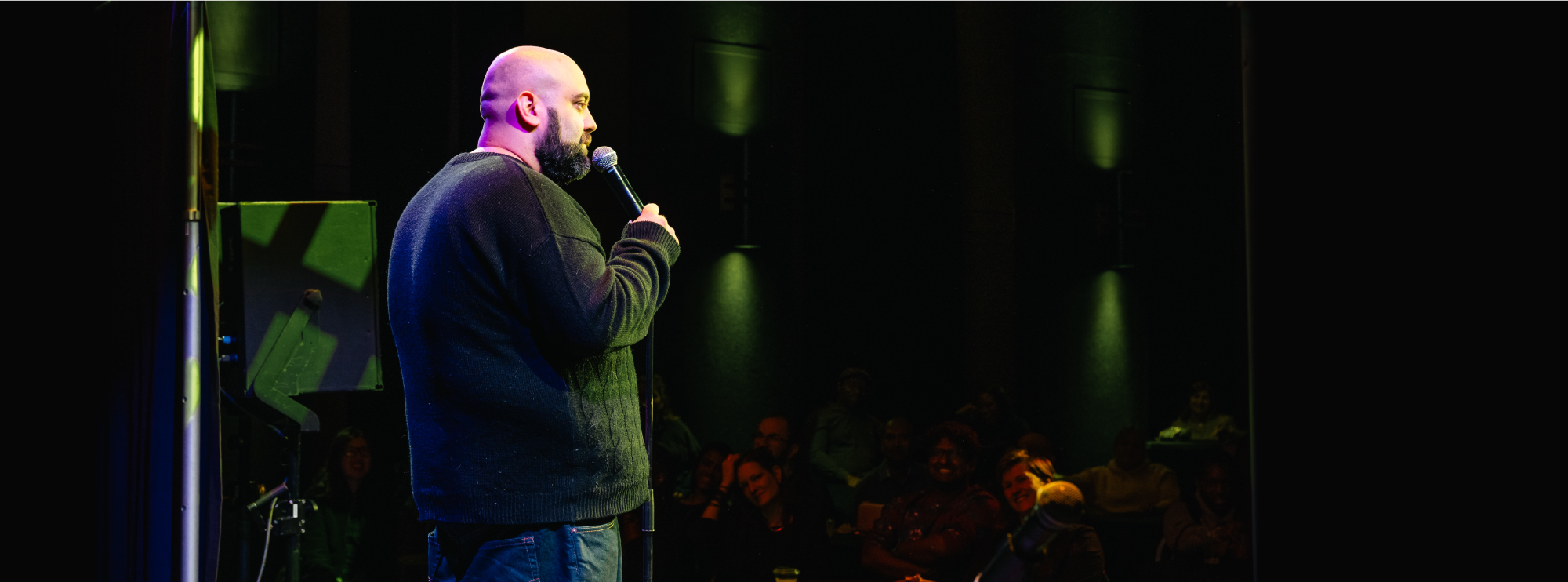 Comedian performing on stage with a microphone, facing an audience seated in a dimly lit venue.