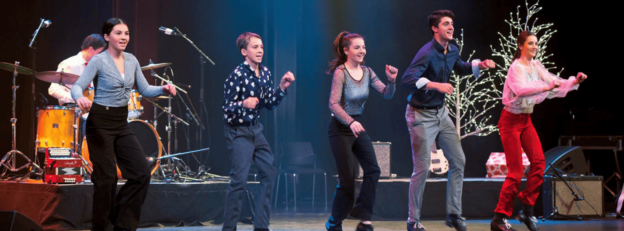 Image of the Leahy family tap dancing on a stage adorned with festive trees and colourful lighting. Three young girls and two young men perform downstage while another musician plays the drums. 