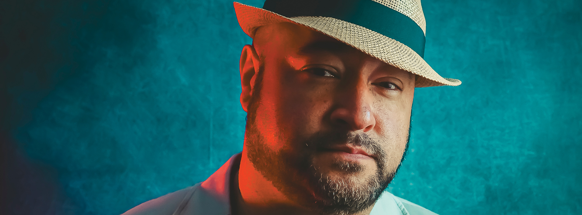 The image features a man wearing a light-colored jacket and a fedora hat with a dark band. His expression is serious and confident, with a subtle red light casting a warm hue on one side of his face, contrasting with the cool, blue-toned background.