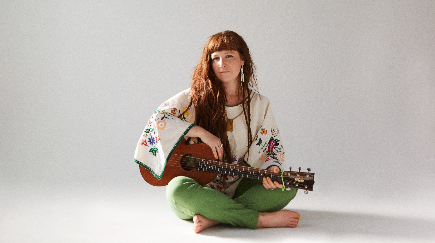 Mimi O'Bonsawin, with long brown hair, seated with a guitar before a white backdrop. She wears floral traditional attire and green pants. 