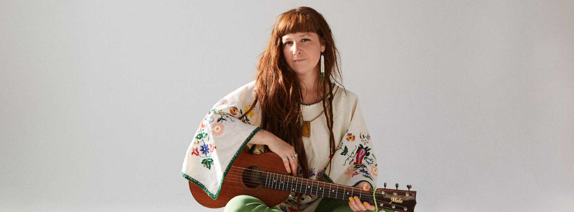 Mimi O'Bonsawin, with long brown hair, seated with a guitar before a white backdrop. She wears floral traditional attire and green pants. 