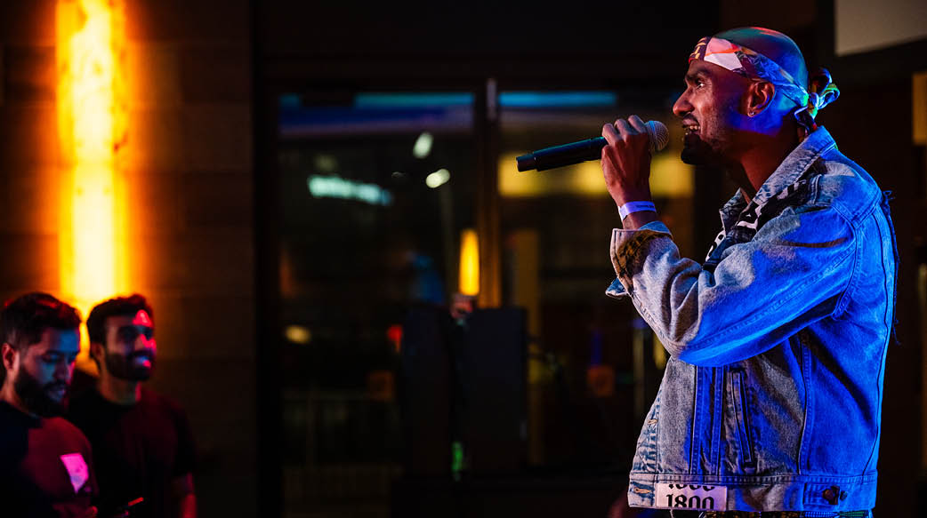 Profile image of an artist performing in front of an audience. He has warm-toned skin and wears a jean jacket with a white bandana on his head. In his left hand, he holds a microphone up towards him. Lighting illuminates the otherwise dark space he is in. People off to the left smile as they him perform. 