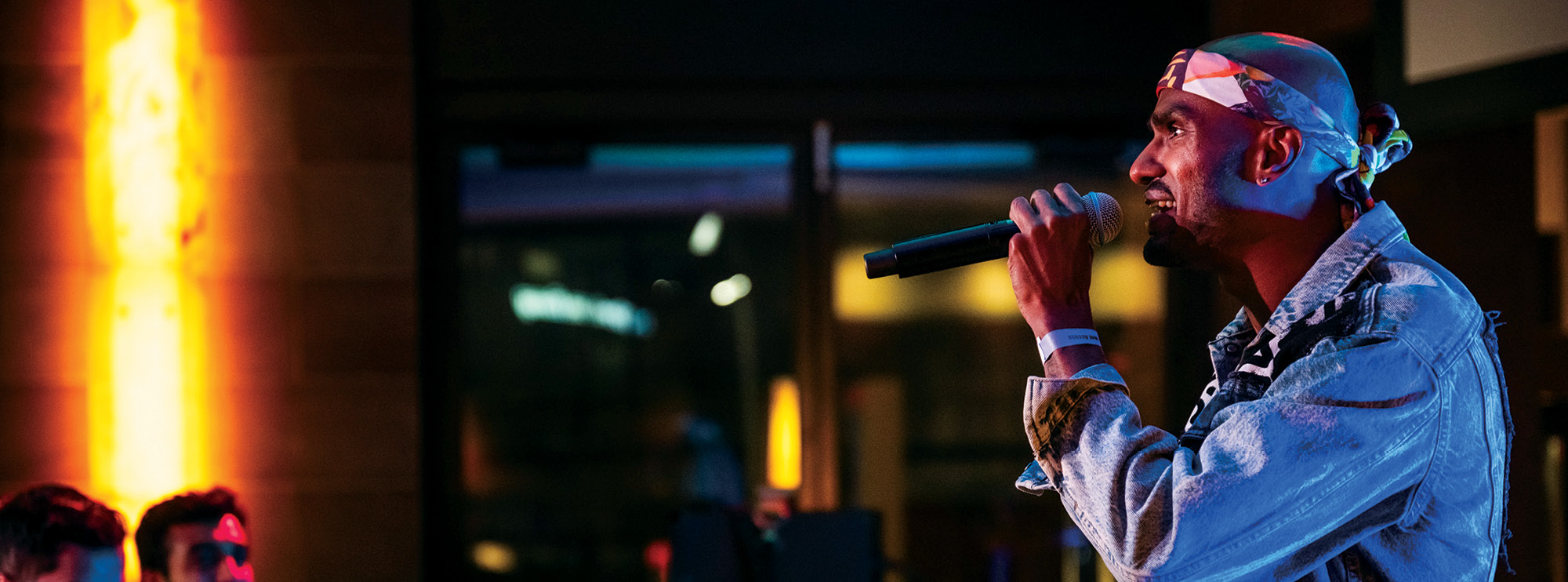 Profile image of an artist performing in front of an audience. He has warm-toned skin and wears a jean jacket with a white bandana on his head. In his left hand, he holds a microphone up towards him. Lighting illuminates the otherwise dark space he is in. People off to the left smile as they him perform. 