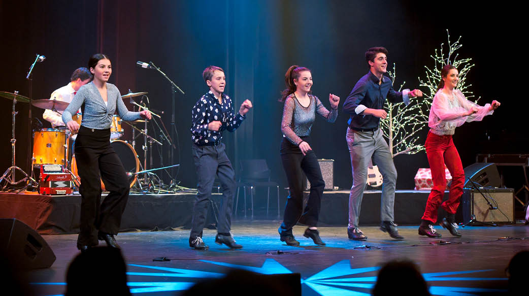 Image of the Leahy family tap dancing on a stage adorned with festive trees and colourful lighting. Three young girls and two young men perform downstage while another musician plays the drums. 