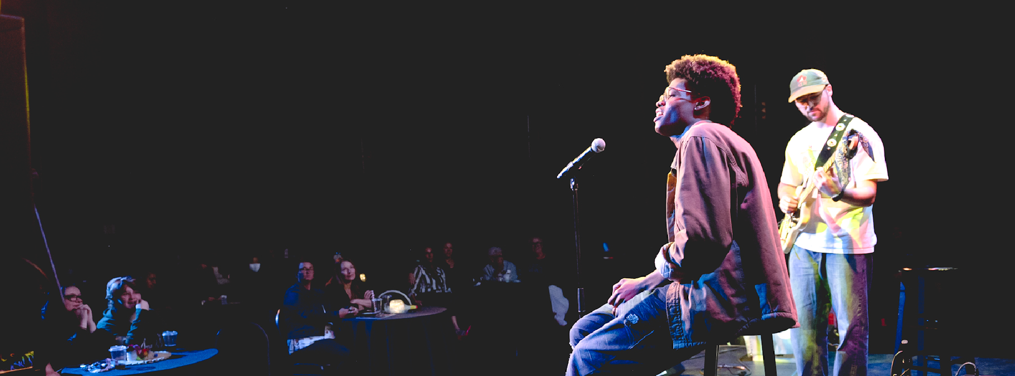 Male vocalist in glasses and denim with guitarist on stage in a dimly lit theater. 