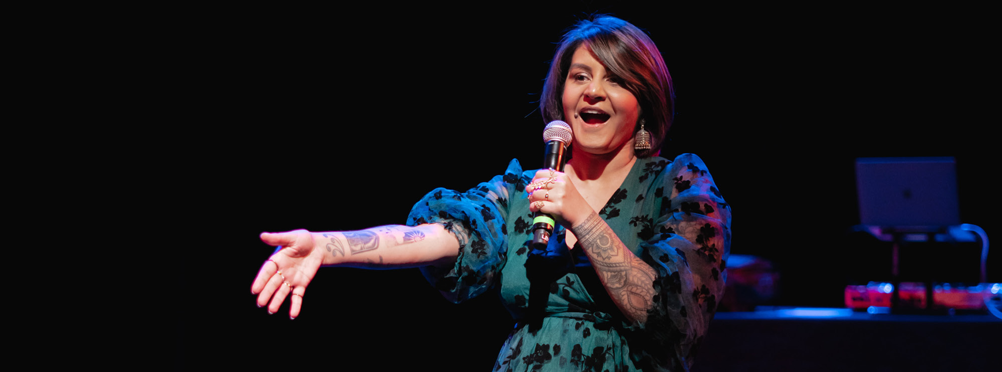 This image captures a lively performance by a woman on stage. She is holding a microphone and appears to be in the midst of delivering an engaging piece, indicated by her animated expression and extended hand gesture. The performer is wearing a stylish teal dress with black floral patterns and has visible tattoos on her arms. She is illuminated by stage lights that create a vibrant and dynamic atmosphere. 