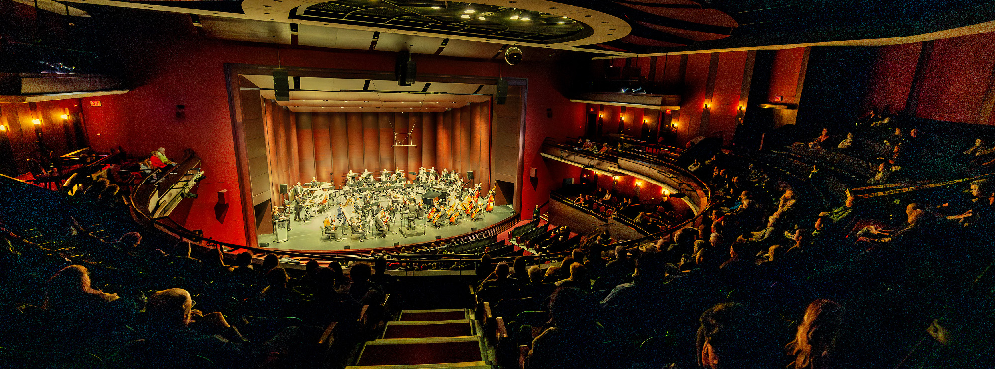 Orchestra playing in a theater with an audience in tiered seating under warm, ambient lighting. 