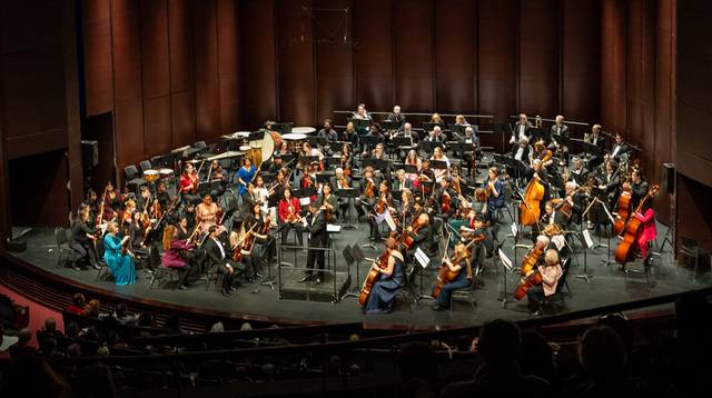 Image of a large orchestra performing on The Rose Mainstage. The musicians are seated in an organized fashion around the stage with music stands before them. They play an array of instruments including the violin, cello and drums. The conductor is stationed at the centre of the stage.  