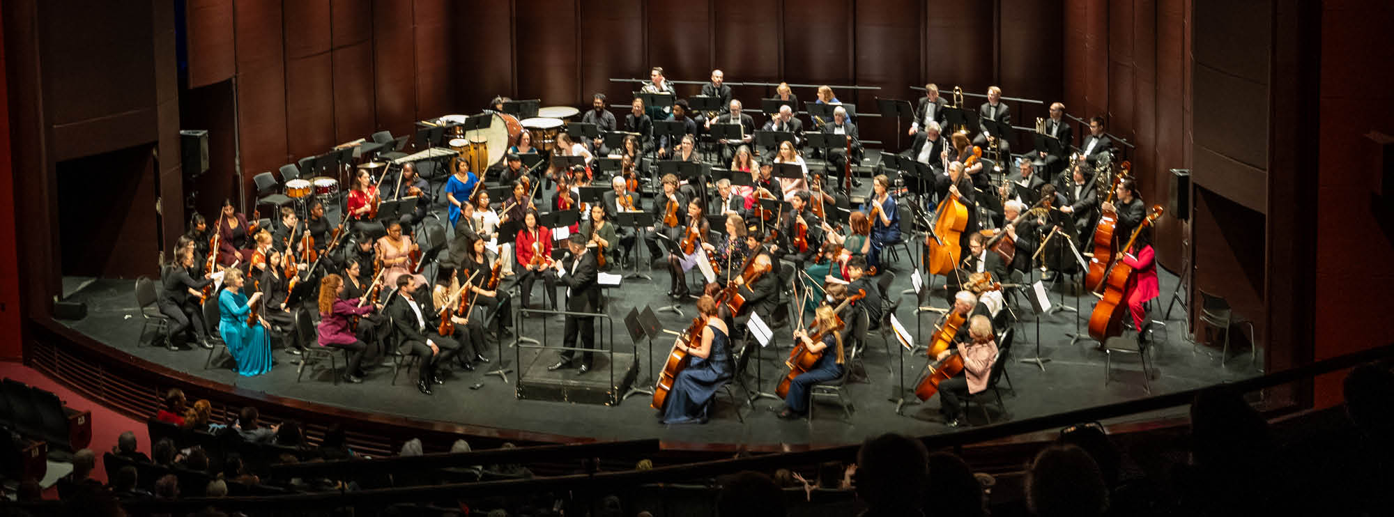 Image of a large orchestra performing on The Rose Mainstage. The musicians are seated in an organized fashion around the stage with music stands before them. They play an array of instruments including the violin, cello and drums. The conductor is stationed at the centre of the stage.  