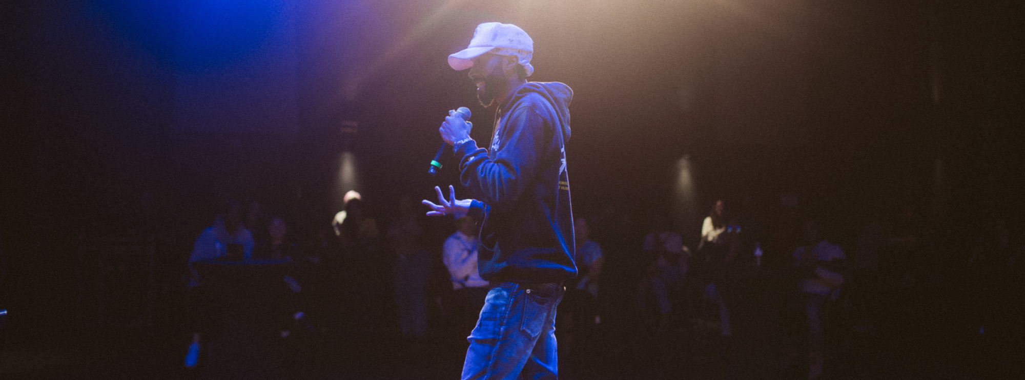 Profile image of the artist TCspades. He walks across the stage, wearing a white baseball cap, a black hooded sweatshirt, and jeans. In his left hand, he holds a microphone, as he smiles and speaks. Beyond him, an audience of people stands, observing him. Lights cast a blue hue on TCspades and the stage.