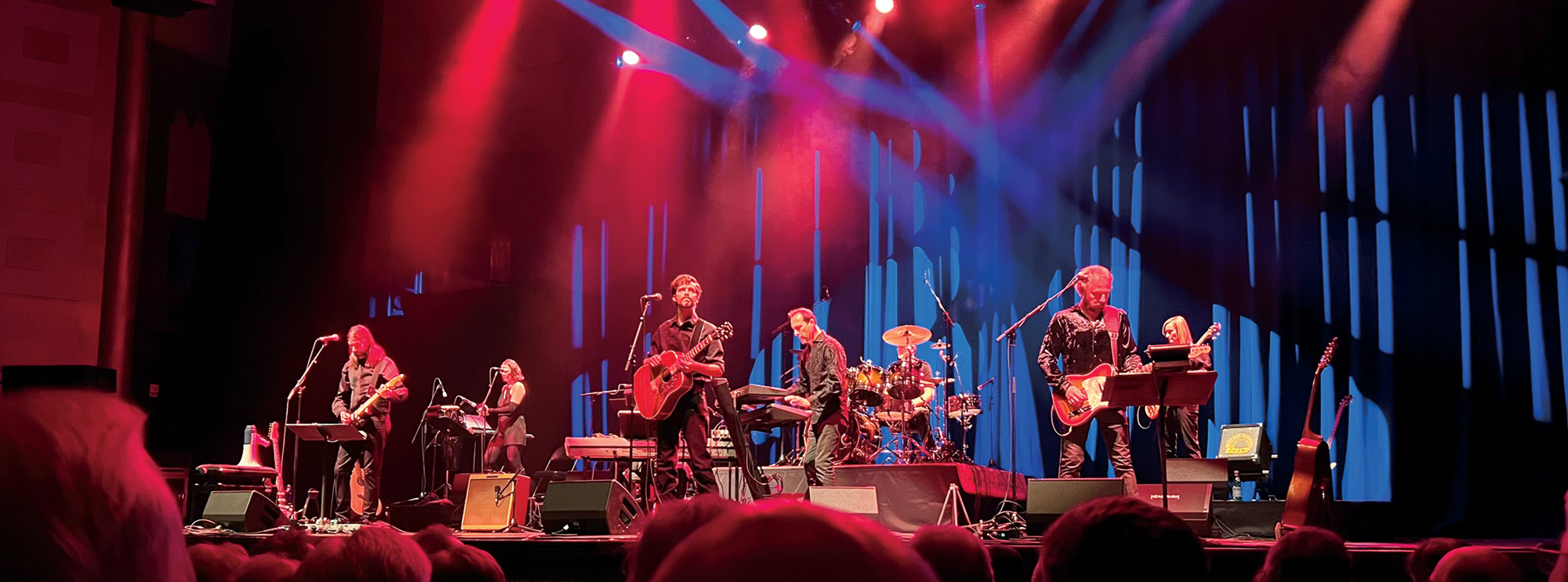 Image of several musicians performing on stage, playing an array of instruments including the guitar, drums, and piano. Speakers, music stands, and wires are scattered across the stage, while blue and red lights illuminate the space. The tops of heads can be seen as the audience observes from below.