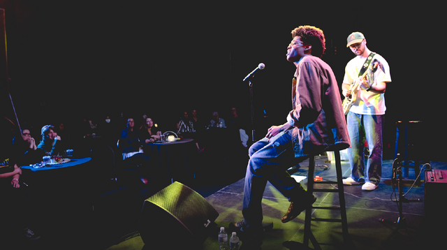 Male vocalist in glasses and denim with guitarist on stage in a dimly lit theater. 