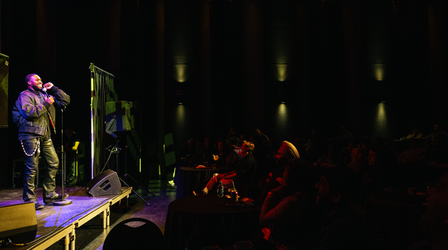 Black male performing on stage to an audience in a dimly lit venue. 