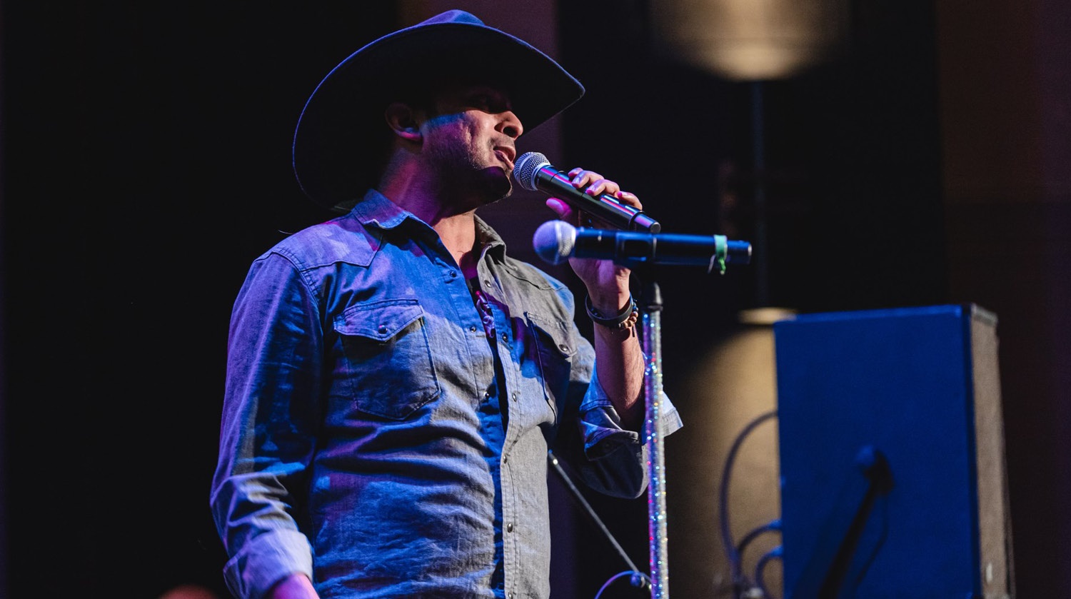 The image captures a male performer wearing a black cowboy hat and a denim shirt, singing into a microphone on stage. The stage is dimly lit with a deep blue and purple ambiance, casting shadows on the background. The performer holds the microphone stand with one hand, and a large speaker and a faint stage light are visible in the background.