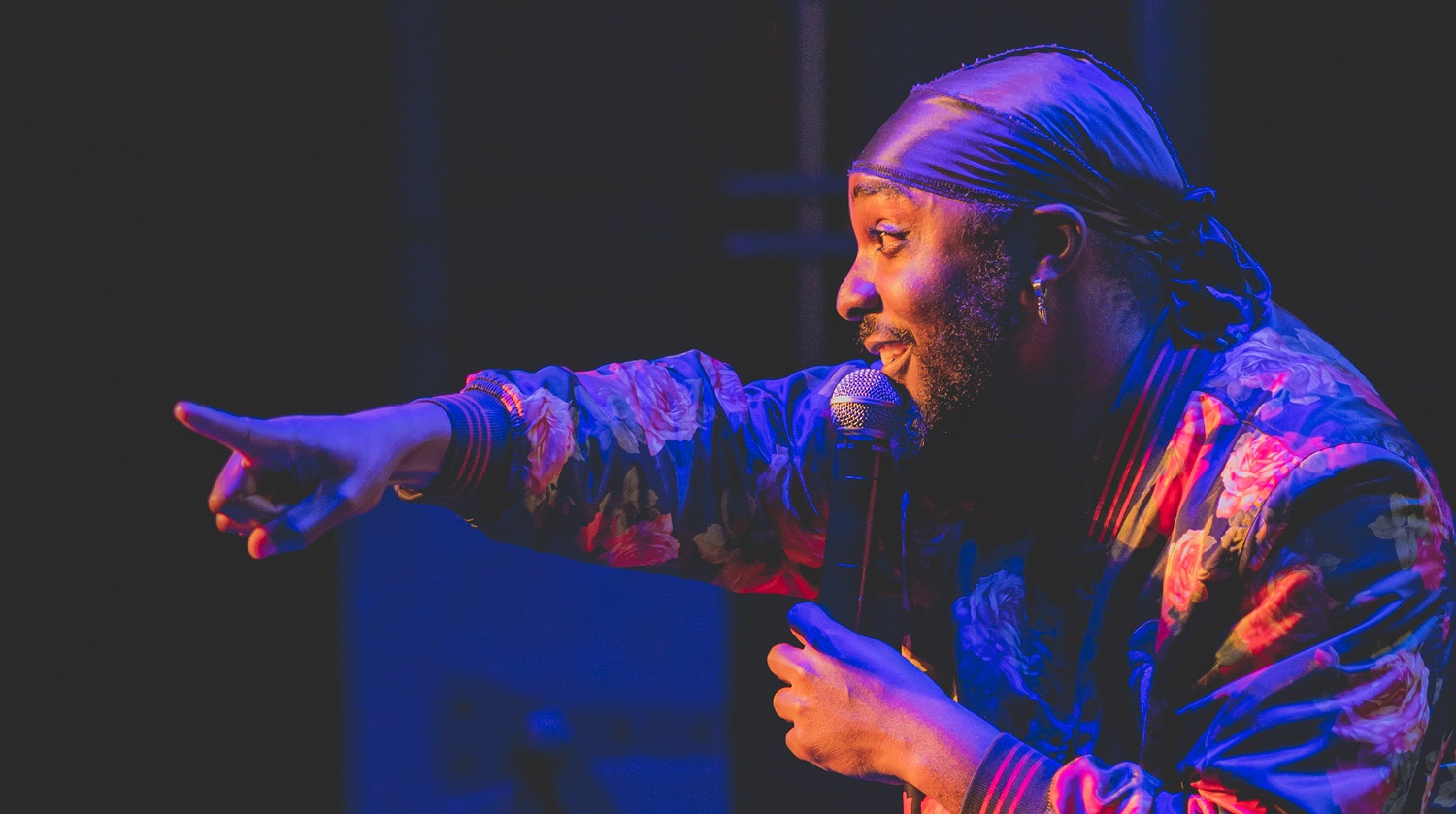 A male comedian stands on stage, holding a microphone and gesturing energetically towards the audience. He wears a colorful floral bomber jacket and a purple headscarf. The stage lighting highlights his face and jacket with hues of purple and orange, adding a dynamic and vibrant atmosphere to the scene. 