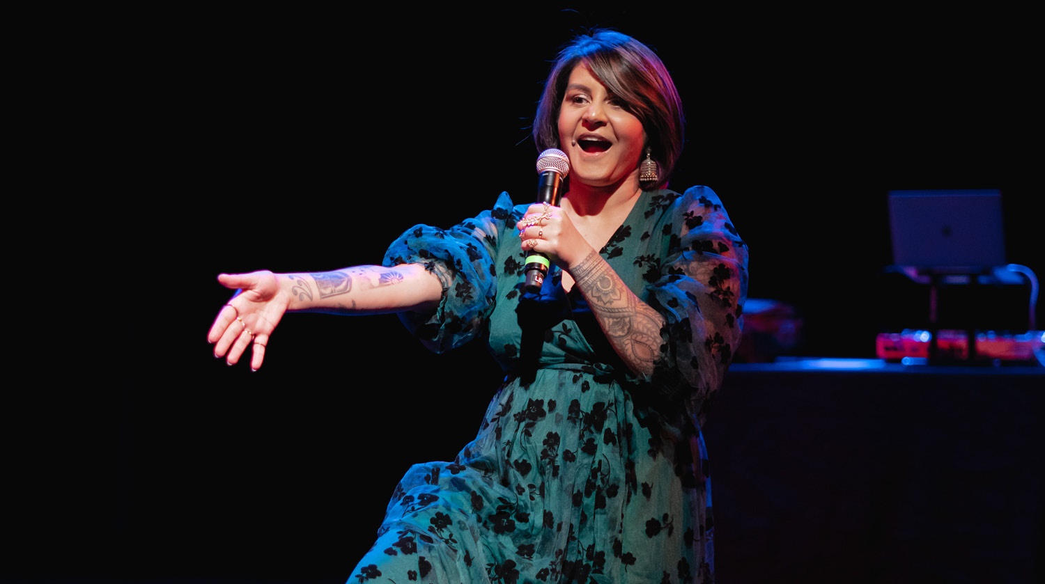 This image captures a lively performance by a woman on stage. She is holding a microphone and appears to be in the midst of delivering an engaging piece, indicated by her animated expression and extended hand gesture. The performer is wearing a stylish teal dress with black floral patterns and has visible tattoos on her arms. She is illuminated by stage lights that create a vibrant and dynamic atmosphere. 