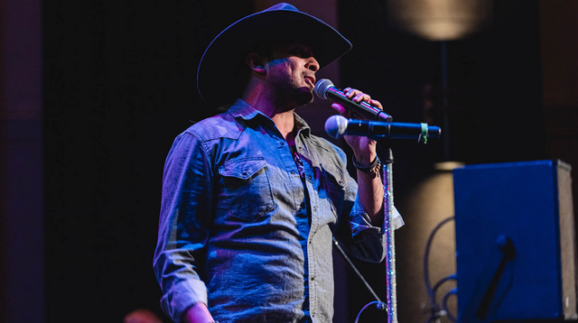 Country singer in denim singing into a microphone on stage, wearing a cowboy hat. 