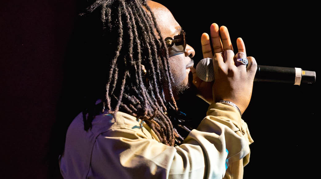 Black male singer with dreadlocks performing against a black backdrop, hand on microphone.