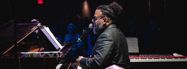 Pianist in a suit performing at a grand piano with a microphone in a blue-lit venue. 