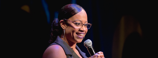 A smiling black woman holding a microphone on stage. She wears glasses and sleeveless top.