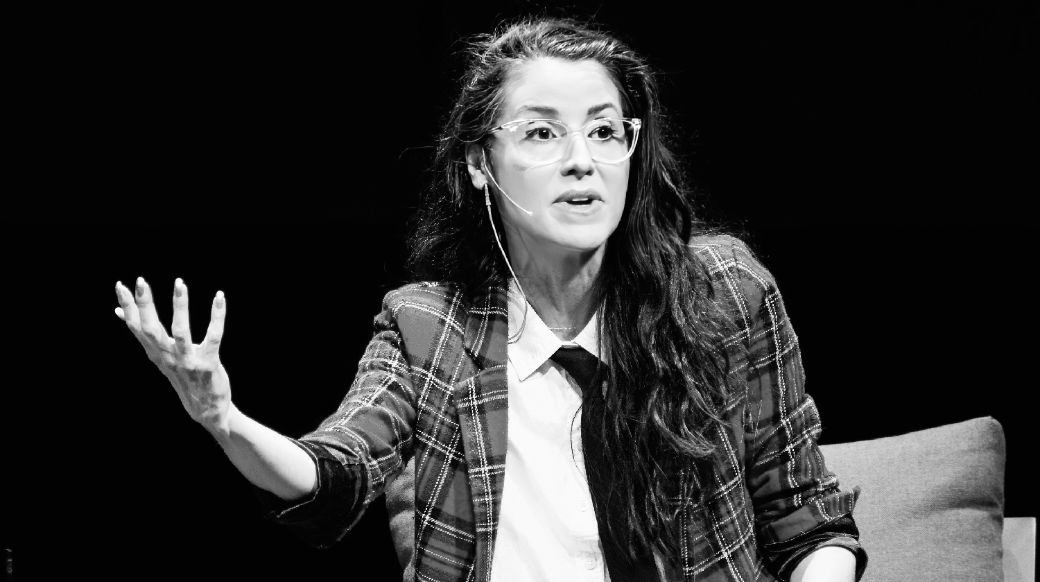 Monochrome image of a woman with glasses gesturing while speaking, wearing a plaid jacket.