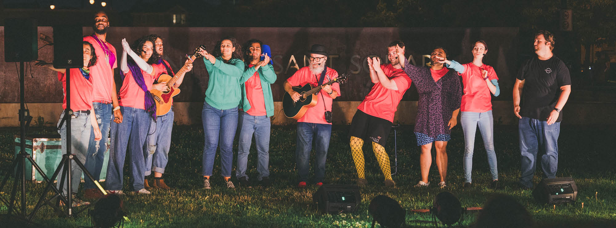 Group of diverse individuals outdoors at night, singing and playing guitar, dressed in red shirts and colorful attire, illuminated by front lights. 