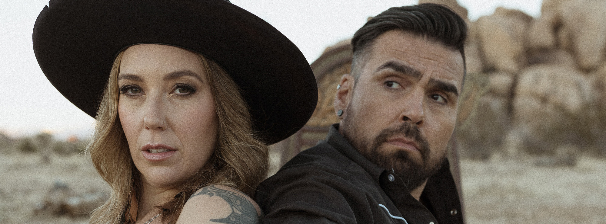 A striking portrait features a woman and a man sitting back-to-back in a rugged outdoor setting, possibly a desert with rocky terrain visible in the background. The woman wears a wide-brimmed black hat and a sleeveless top. The man has a rugged look with short hair, a beard, and is dressed in a dark shirt with white piping.