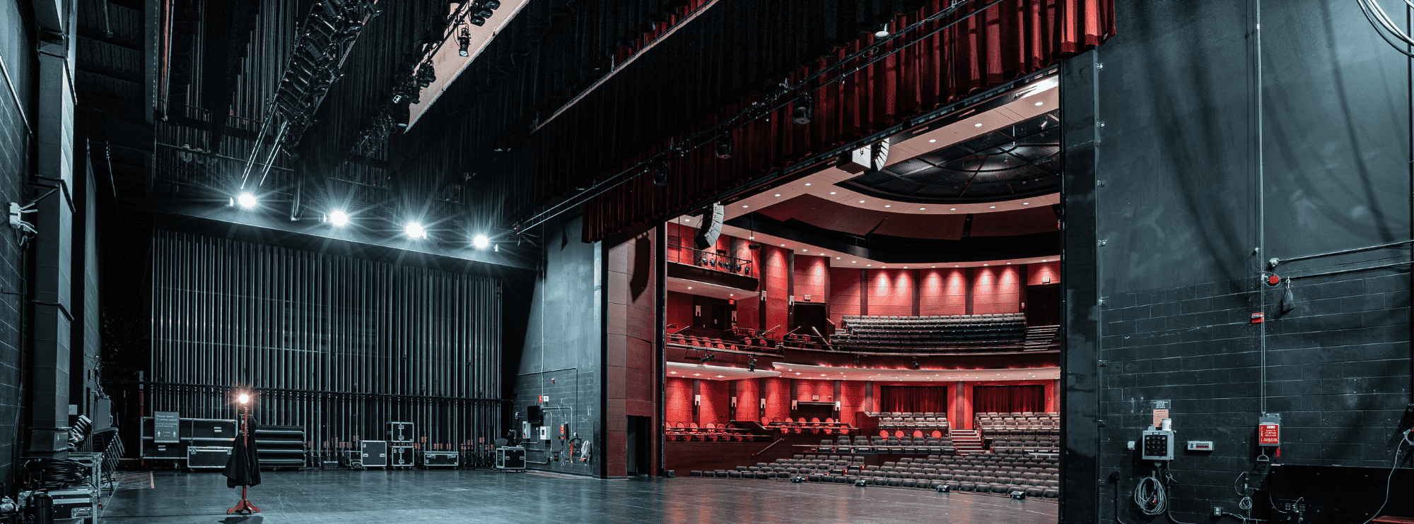 View from backstage of a theater, showing the stage, equipment, and empty auditorium with red seating and walls.