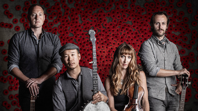 Image of four musicians clad in grey attire set against a backdrop of poppies. Positioned on either side, the musicians stand facing forward, their hands resting on guitars. In the center, one musician holds a banjo, while the another has long brown hair and holds a violin.