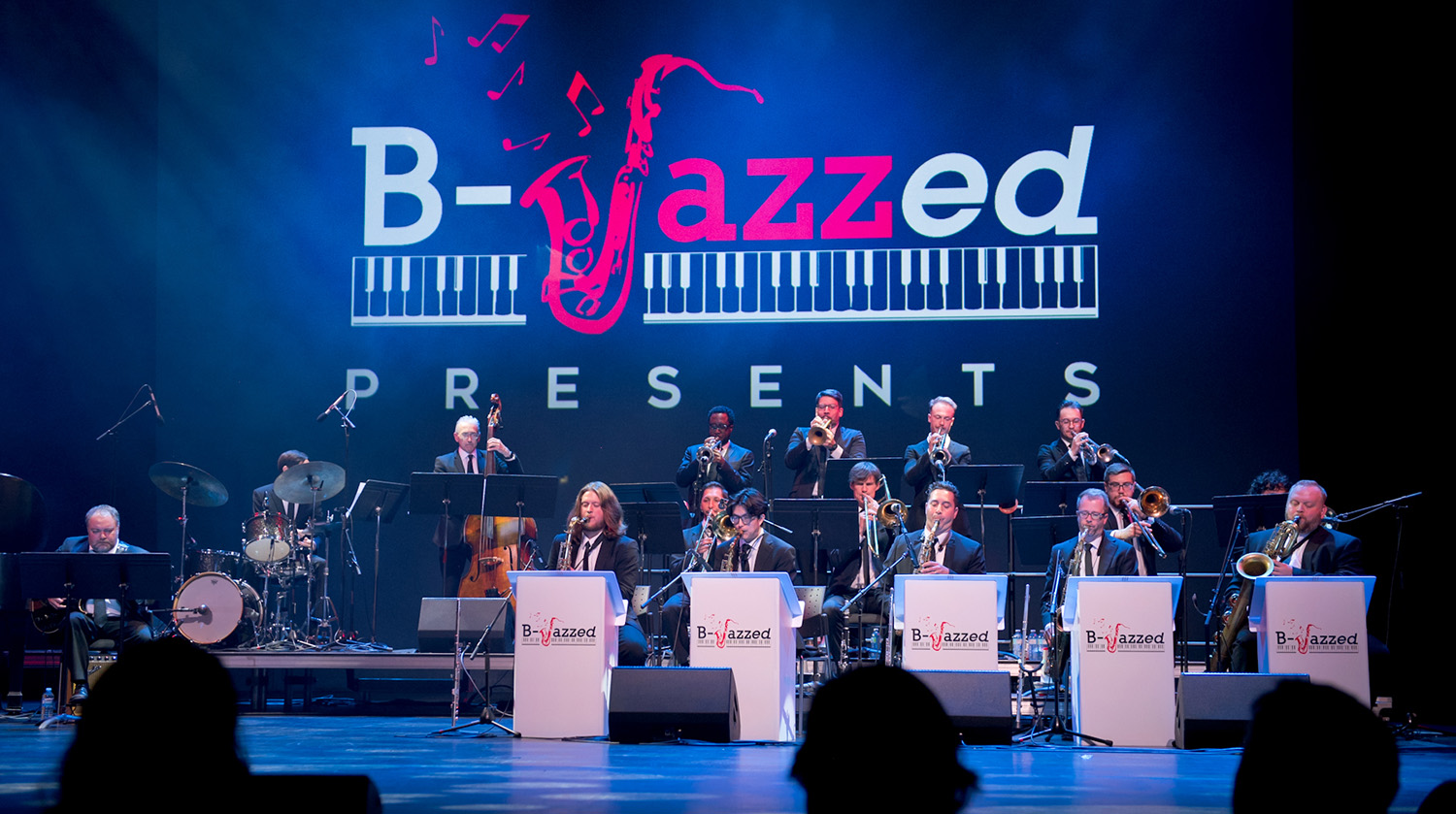 A big band jazz ensemble performs on stage in front of a large screen displaying the "B-Jazzed Presents" logo, with musicians playing brass, woodwind, and rhythm instruments under blue stage lighting.