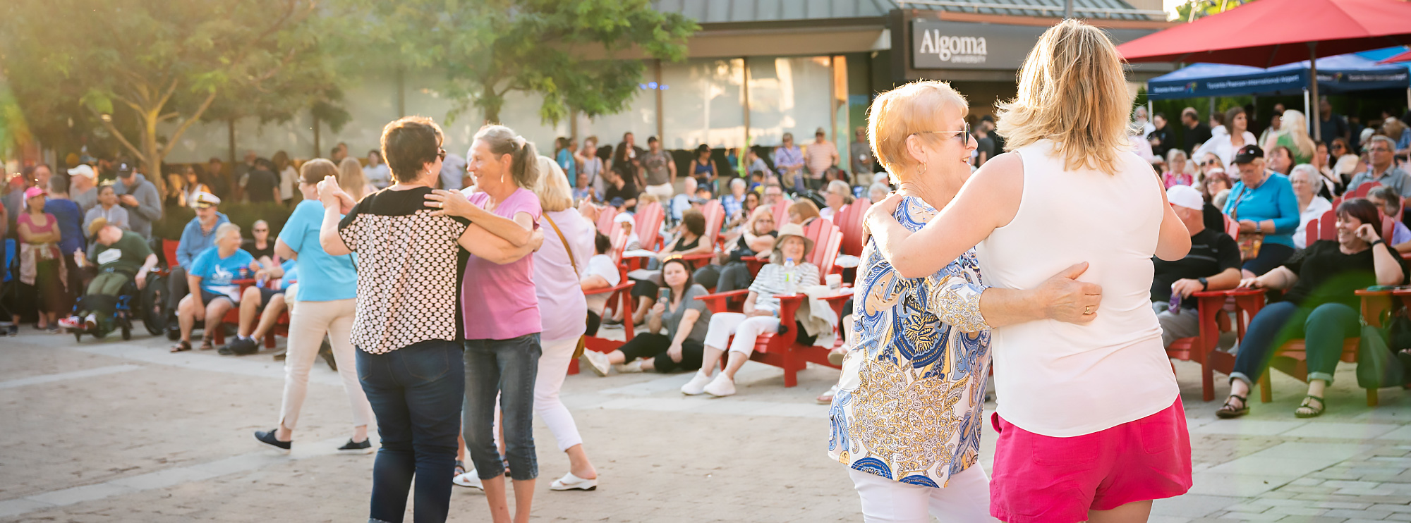 Two pairs of women dance together in an outdoor plaza, smiling and enjoying the music. Behind them, a crowd sits on red chairs and stands under umbrellas, watching the festivities. The atmosphere is lively and sunlit, suggesting a cheerful community event.