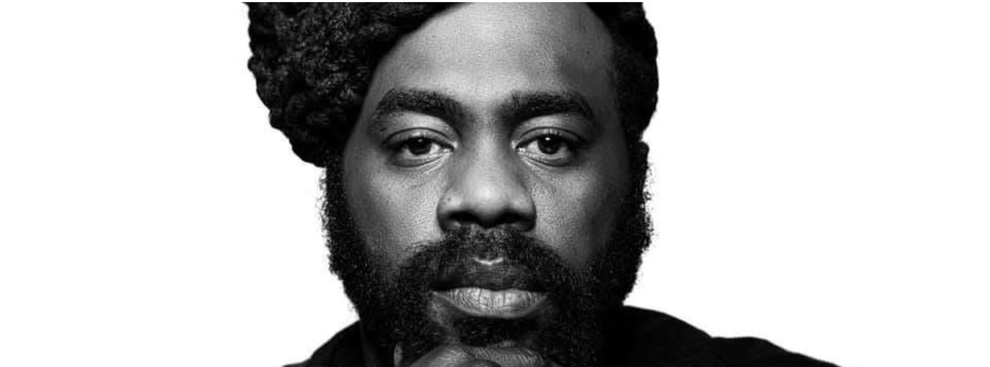 Black and white close-up portrait of a man with a serious expression, a full beard, and thick hair styled in a wrapped bun. He rests his chin lightly on his hand, gazing directly at the camera against a white background