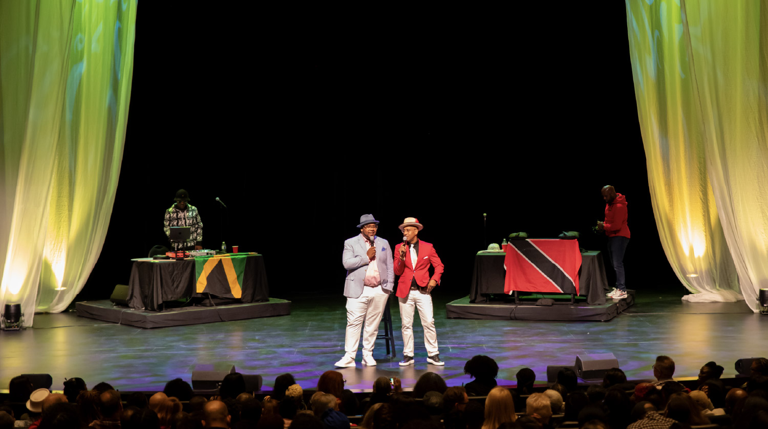 Two men stand at centre stage in front of a live audience, each holding a microphone. One wears a light blue blazer and fedora, the other a red blazer and matching hat. On either side of them, DJs stand behind tables draped with the Jamaican and Trinidadian flags. The stage is lit with soft yellow and green lighting, with tall, flowing curtains framing the scene.