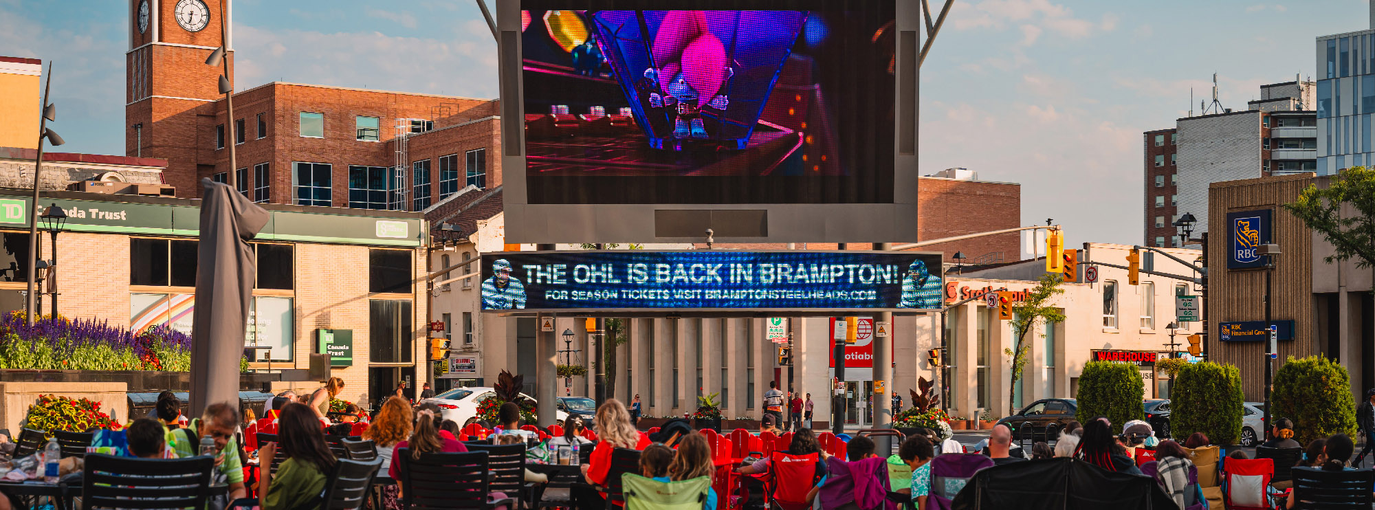 A crowd of people seated in red lawn chairs watches a large outdoor screen in downtown Brampton during a public event. The digital billboard below the screen reads “THE OHL IS BACK IN BRAMPTON!” promoting local hockey. Surrounding buildings include TD, RBC, and Scotiabank branches, with the Brampton clock tower visible in the background.