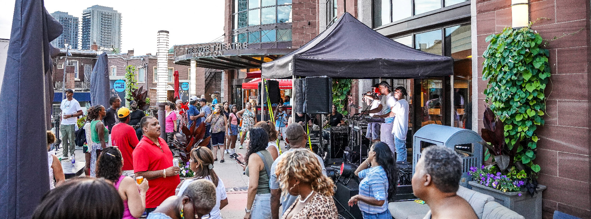 A lively crowd gathers outside The Rose Theatre in Brampton for a patio performance. Under a black tent, a DJ and emcee perform while people dance, mingle, and enjoy the upbeat atmosphere. Tall buildings and greenery frame the urban summer scene.