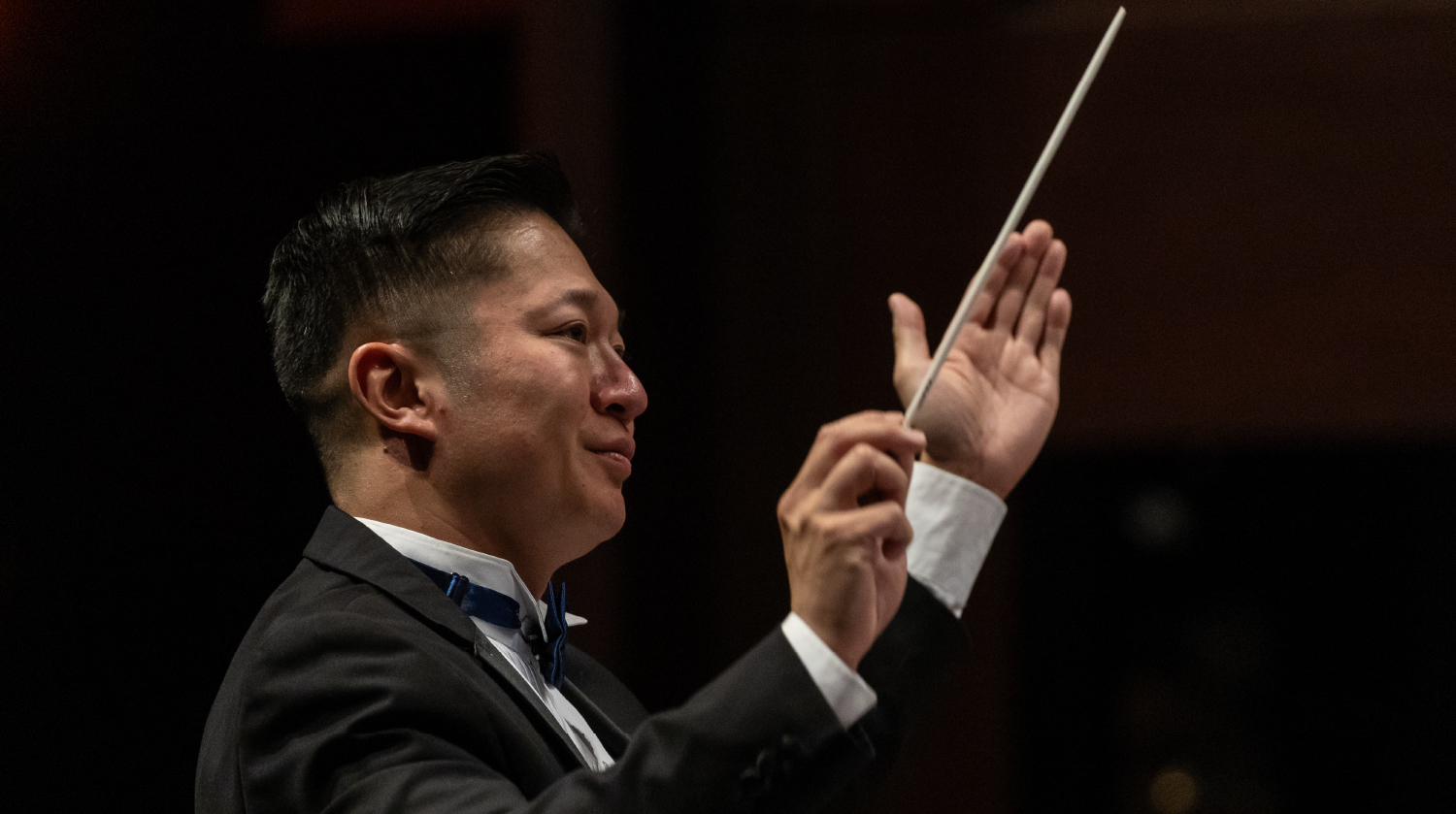 A conductor in a black tuxedo and blue bow tie leads an orchestra with precision, holding a baton in one hand while gesturing expressively. Focused lighting highlights his concentrated expression against a dark background.
