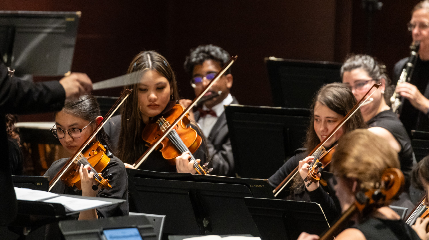 A group of violinists performs with focus and precision as a conductor leads them. Dressed in black, they play under warm stage lighting, with woodwind musicians visible in the background.