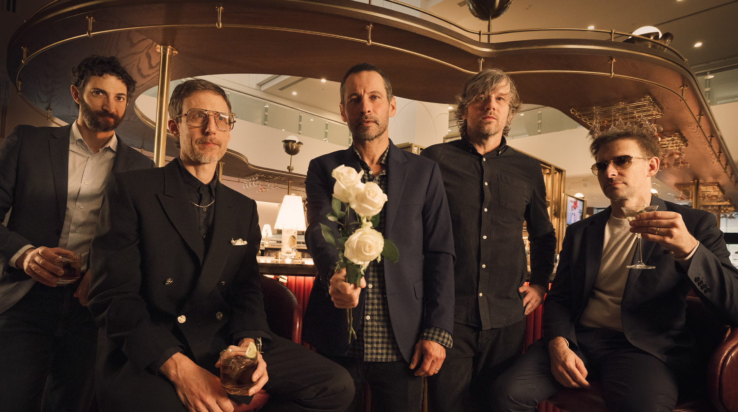 Five men pose together inside an upscale lounge or hotel bar. They are dressed in stylish suits and semi-formal outfits, holding drinks in cocktail glasses. The man in the centre holds a bouquet of white roses and gazes directly at the camera. Warm ambient lighting, modern decor, and a curved wooden bar with brass fixtures create a sophisticated atmosphere in the background. The group appears to be a band or ensemble, exuding a moody yet polished vibe.